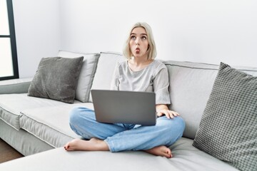 Young caucasian woman using laptop at home sitting on the sofa making fish face with lips, crazy and comical gesture. funny expression.