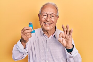 Senior man with grey hair holding medical asthma inhaler doing ok sign with fingers, smiling friendly gesturing excellent symbol - obrazy, fototapety, plakaty