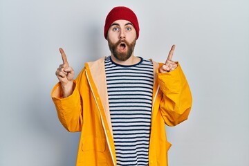 Caucasian man with beard wearing yellow raincoat amazed and surprised looking up and pointing with fingers and raised arms.