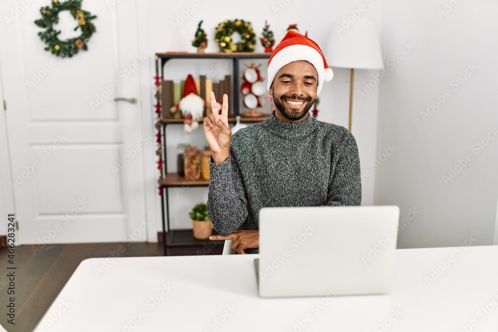 Canvas Prints young hispanic man with beard wearing christmas hat using laptop smiling with happy face winking at 