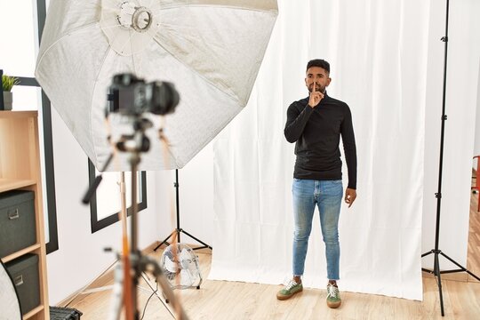 Young hispanic man with beard posing as model at photography studio asking to be quiet with finger on lips. silence and secret concept.