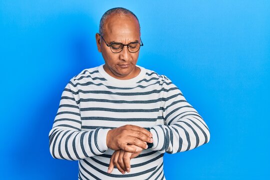 Middle age latin man wearing casual clothes and glasses checking the time on wrist watch, relaxed and confident