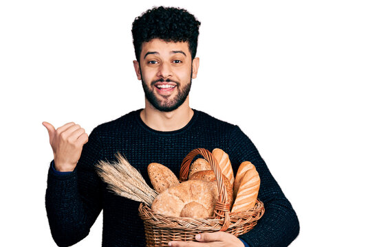 Young arab man with beard holding wicker basket with bread pointing thumb up to the side smiling happy with open mouth