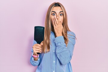 Beautiful hispanic woman holding hairbrush covering mouth with hand, shocked and afraid for...