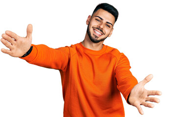 Young hispanic man with beard wearing casual orange sweater looking at the camera smiling with open arms for hug. cheerful expression embracing happiness. - obrazy, fototapety, plakaty