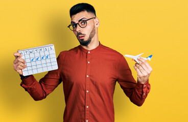 Young hispanic man with beard holding plane toy and travel calendar in shock face, looking...