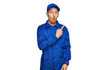 Bald man with beard wearing builder jumpsuit uniform surprised pointing with finger to the side, open mouth amazed expression.