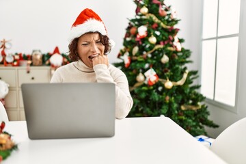 Middle age hispanic woman using laptop sitting by christmas tree looking stressed and nervous with hands on mouth biting nails. anxiety problem.