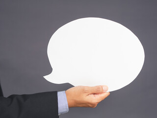 Business and speech bubble concept. Businessman in suit hand holding an empty speech bubble paper while standing on gray background in the studio. Close-up photo