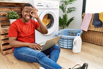 Young african man doing laundry and using computer doing ok gesture with hand smiling, eye looking through fingers with happy face.