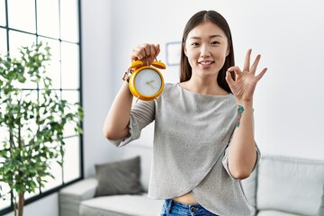 Young asian woman holding alarm clock doing ok sign with fingers, smiling friendly gesturing excellent symbol