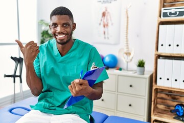 Young african american man working at pain recovery clinic pointing to the back behind with hand and thumbs up, smiling confident