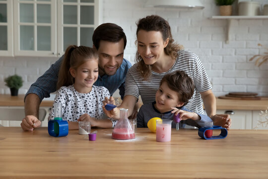 Happy young caring couple parents showing chemical experiments to interested cheerful little kids son daughter in modern kitchen, enjoying learning together with game, education with entertainment.