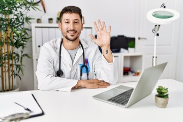 Young doctor working at the clinic using computer laptop showing and pointing up with fingers number five while smiling confident and happy.