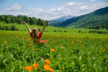 A young blond girl wearing red dress sitting on a green lawn, and looking downhill over valley above cloud in mountains. Back view. Green valley high on the mountains