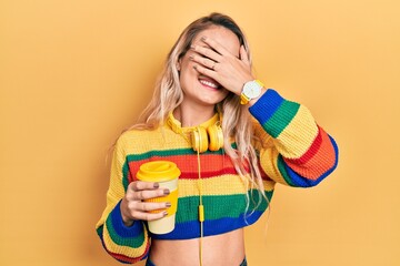 Beautiful young blonde woman drinking cup of coffee wearing headphones smiling and laughing with hand on face covering eyes for surprise. blind concept.