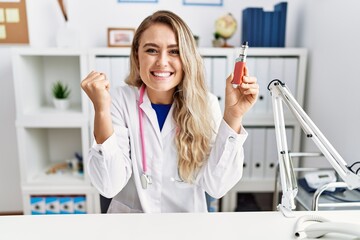 Young beautiful doctor woman holding electronic cigarette at the clinic screaming proud, celebrating victory and success very excited with raised arms