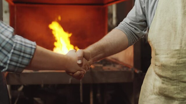 Close up shot of two blacksmiths shaking hands and bumping fists while standing by forge in workshop