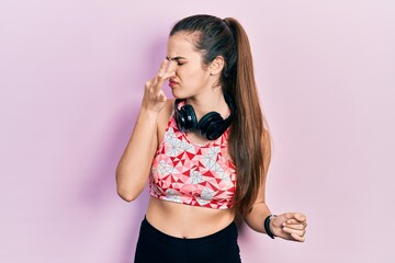 Young brunette teenager wearing gym clothes and using headphones smelling something stinky and disgusting, intolerable smell, holding breath with fingers on nose. bad smell