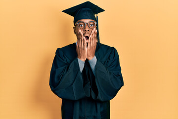 Young african american man wearing graduation cap and ceremony robe afraid and shocked, surprise and amazed expression with hands on face