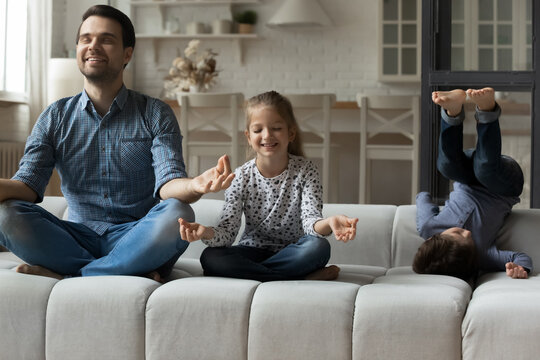 Happy young father sitting on cozy sofa in lotus position with small kid daughter, teaching making yoga breathing exercises or asanas, enjoying domestic hobby activity while small son having fun. - Powered by Adobe
