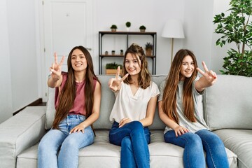 Group of three hispanic girls sitting on the sofa at home smiling looking to the camera showing fingers doing victory sign. number two.