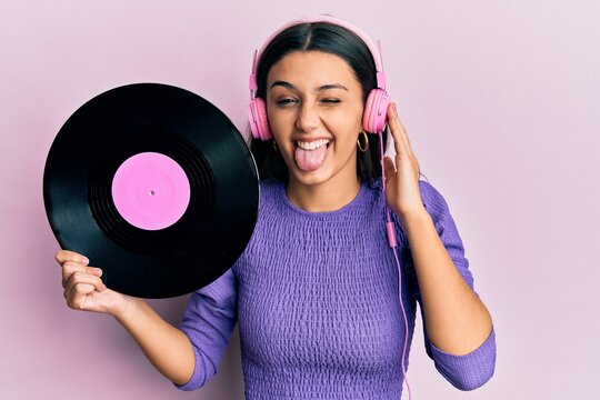 Young hispanic woman using headphones holding vinyl disc sticking tongue out happy with funny expression.
