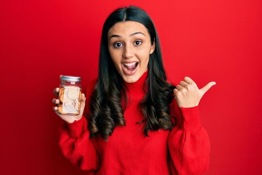 Young hispanic woman holding salty biscuits jar pointing thumb up to the side smiling happy with open mouth