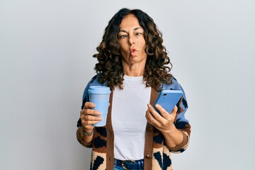 Middle age hispanic woman drinking a cup of coffee and looking at the smartphone screen making fish face with mouth and squinting eyes, crazy and comical.