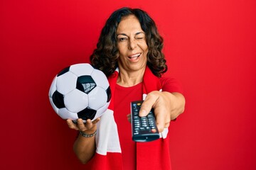 Middle age hispanic woman football hooligan holding ball using tv control winking looking at the...
