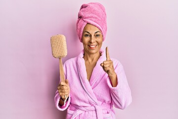 Middle age hispanic woman wearing bathrobe holding sponge smiling with an idea or question pointing finger with happy face, number one