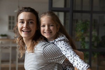 Portrait of beautiful young mother giving piggyback ride to little child daughter, happy millennial mum and cute small kid girl having fun, entertaining together on weekend at home, looking at camera.