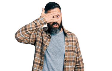 Hispanic man with beard wearing casual shirt peeking in shock covering face and eyes with hand, looking through fingers with embarrassed expression.