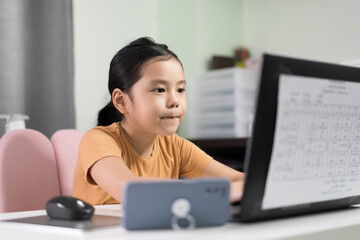 Asian child student or kid girl purse lips to enjoy diligent studying on computer laptop and smartphone by video call study online class or person learn from home school to play notebook for education