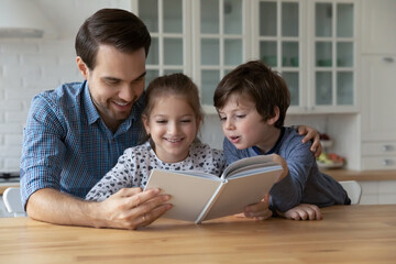 Happy young father reading paper book to little adorable kids son daughter, telling interesting...