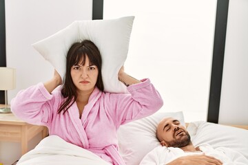 Hispanic woman angry with pillow on his ears while man snoring on the bed.