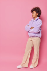 Obraz na płótnie Canvas Full length shot of thoughtful self confident woman with curly hair stands indoor with arms folded wears fur jacket and bell bottomed trousers isolated over pink background blank copy space.