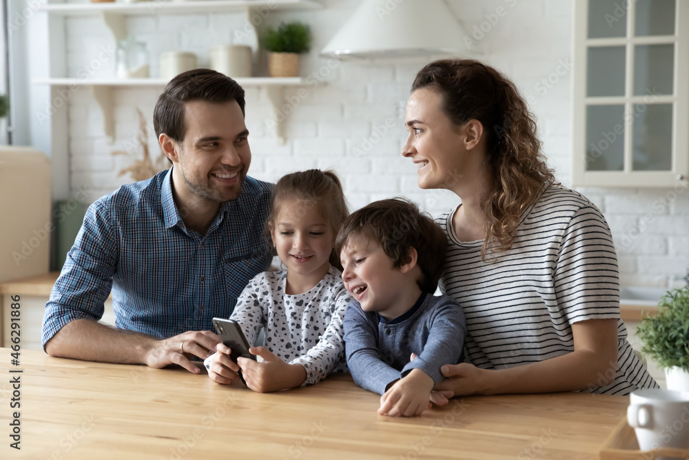 Wall mural Happy friendly young couple parents using cellphone with small kids, having fun entertaining online together, playing mobile games or watching funny photo video content in social network at home.