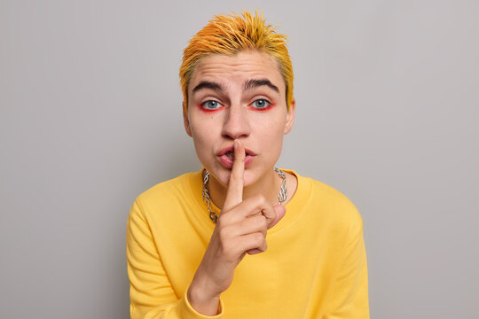Serious blue eyed woman punk with trendy yellow hairstyle bright colorful makeup makes silence gesture shushing indoor wears casual jumper poses against grey background. Hush be quiet please