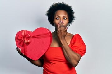 Young african american woman holding valentine gift covering mouth with hand, shocked and afraid...