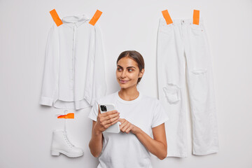 Pleased woman uses mobile phone for selling her used clothes online surrounded by white shirt trousers and boot plastered to background looks happily away. People clothes and technology concept