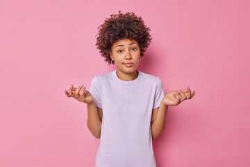 Perplexed indecisive young curly haired woman spreads palms sideways has doubtful expression expresses uncertainty wears casual t shirt isoated over pink background stands unaware knows nothing - obrazy, fototapety, plakaty