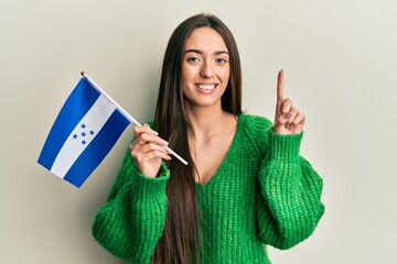 Young hispanic girl holding honduras flag smiling with an idea or question pointing finger with...