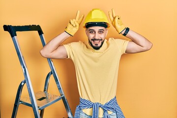 Handsome man with beard by construction stairs wearing hardhat posing funny and crazy with fingers...