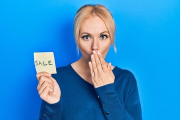 Young blonde woman holding sale word reminder covering mouth with hand, shocked and afraid for...