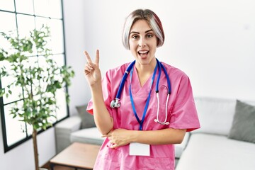 Young beautiful woman wearing doctor uniform and stethoscope smiling with happy face winking at the...