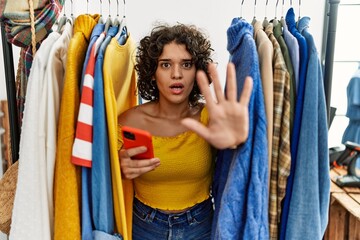Young hispanic woman searching clothes on clothing rack using smartphone doing stop gesture with hands palms, angry and frustration expression