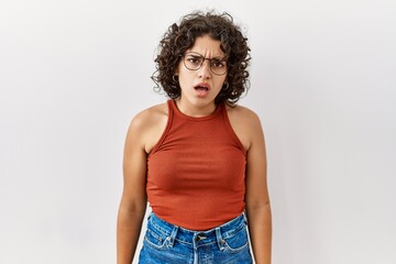 Young hispanic woman wearing glasses standing over isolated background in shock face, looking skeptical and sarcastic, surprised with open mouth