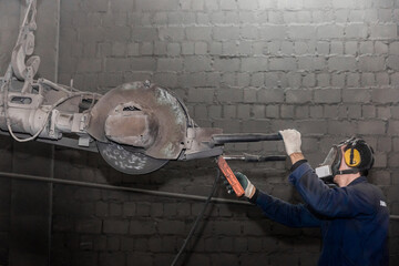 A man in work clothes and a respirator, controls heavy equipment with grinding stone to clean reinforced concrete structures in a dirty shop of an industrial plant