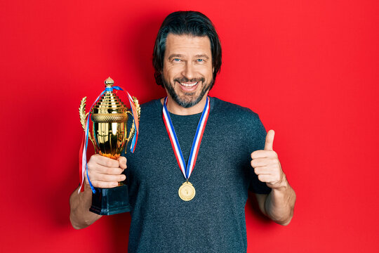 Middle age caucasian man holding champion trophy wearing medals smiling happy and positive, thumb up doing excellent and approval sign